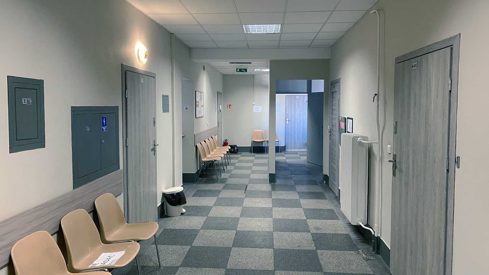 Jasny korytarz w placówce medycznej z ławkami i drzwiami prowadzącymi do innych pomieszczeń.