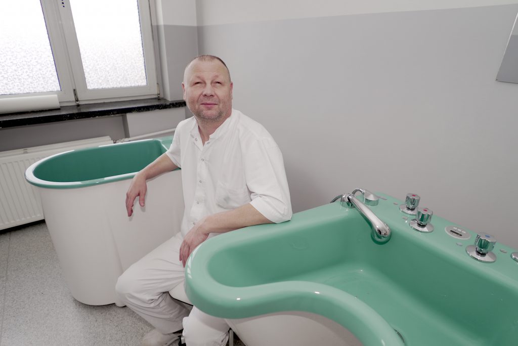 Mężczyzna w białym ubraniu medycznym siedzi obok zielonej wanny w jasnym pomieszczeniu.