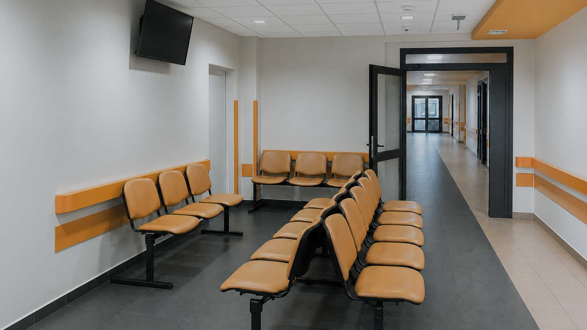 Jasny korytarz w placówce medycznej z pomarańczowymi krzesłami i drzwiami prowadzącymi do innych pomieszczeń.